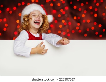 A Portrait Of A Laughing Cute Little Girl In Santa Hat Looking Out From Behind Empty Blank With Her Hands On It, Isolated Against Christmas Glitter Close Up. Holidays Concept. Invitation. Funny Kids. 