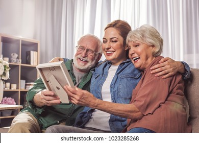 Portrait Of Laughing Grandparents And Smiling Young Woman Looking At Photo In Room. Remembrance Concept