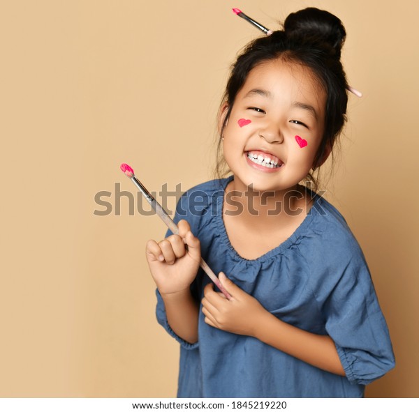 Portrait of\
laughing happy brunette chinese girl with painted red hearts on\
cheeks with brush in hair and hand over yellow wall background.\
Trendy children fashion, asian outfit\
concept