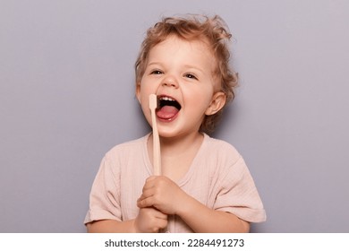 Retrato de una niña alegre y risible con pelo rubio y rizado lavándose los dientes, cuidando la higiene dental aislada sobre fondo gris.