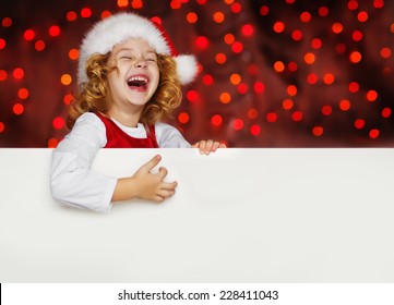 A Portrait Of A Laughing Sweet Little Curly Girl In Santa Hat Looking Out From Behind Empty Blank, Isolated Against Christmas Glitter Close Up. Holidays Concept. Invitation. Funny Kids.