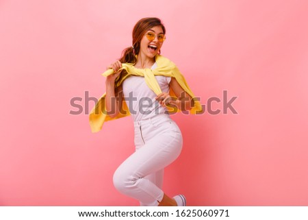  portrait of a laughing woman jumping over pink background. Looking at camera. Beautiful young woman in sunglasses,white shirt,white jeans posing, jumping. Copy space.