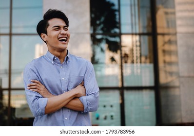 Portrait Of Laughing Young Asian Businessman In The City. Crossed Arms And Looking Away. A Happy Friendly Man