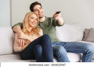 Portrait of a laughing young couple hugging while sitting together on a sofa at home and watching TV