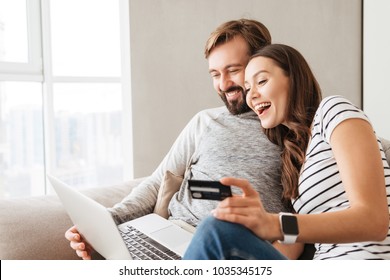 Portrait of a laughing young couple shopping online while sitting on a couch with credit card and laptop computer