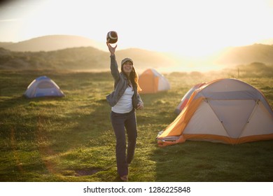 Portrait Of A Laughing Young Woman Throwing A Football In A Campsite At Sunset.