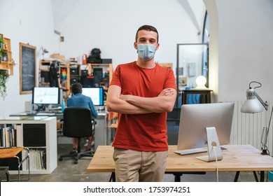 Portrait Of Leader Of A Startup In The Office With Medical Mask For Protection And Prevention Contagions From Coronavirus, Covid-19, Behind Him The Other Workstations With Colleagues At Computer