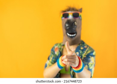 Portrait Of Lesbian Female Wearing Crazy Horse Animal Mask And LGBTQ+ Pride Rainbow Wristband And Sunglasses Pointing At The Camera Isolated On Orange Background