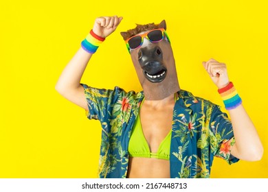 Portrait Of Lesbian Woman Dancing Wearing Crazy Horse Animal Mask LGBTQ+ Pride Rainbow Wristband And Sunglasses Pointing At The Camera Isolated On Yellow Background