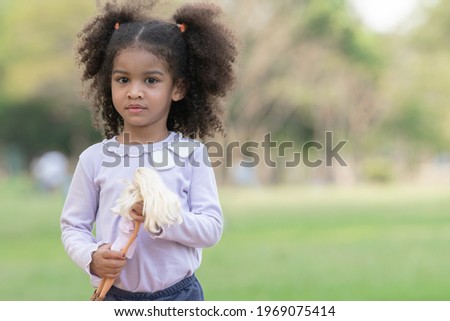 Portrait of little African kid girl with twin tails hair smiling and holding a doll in her hands at green park