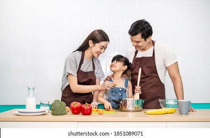 Portrait Of Little Asian Girl And Parents Cooking Food With Pan In The Kitchen. Happy Asian Family With Father Mother Daughter, Mother's Day, Healthcare Cooking Plant Based Food Concept