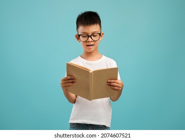 Portrait Of Little Asian Schoolboy In Eyeglasses Reading Book While Standing Isolated Over Blue Studio Background, Nerdy Preteen Korean Male Kid Enjoying Study And Learning Process, Copy Space