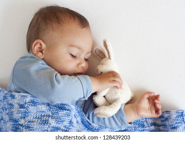 Portrait Of A Little Baby Boy Sleeping Under A Blue Blanket Holding A White Soft Toy Rabbit.