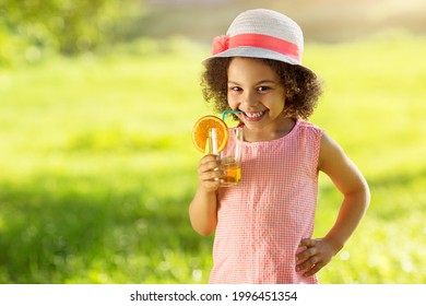 Portrait of little Black girl drinking orange juice in a glass with straw at outdoor park. Child in summer hat suck from the straw a fresh juice