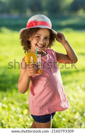 Portrait of little Black girl drinking orange juice in a glass with straw at outdoor park. Child in summer hat suck from the straw a fresh juice
