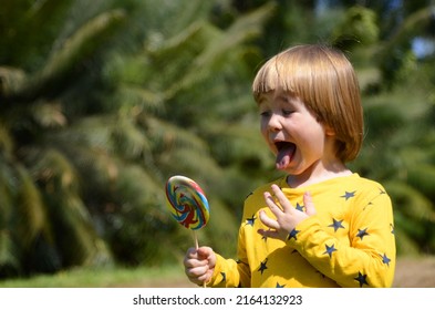 Portrait Of A Little Boy, Background Of Palm Branches. The Child Eats A Big Lolipop. Concept: Vacation, Children's Entertainment, Amusement Park, Family Tropical Resort. Boy Showing Tongue