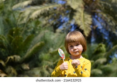 Portrait Of A Little Boy, Background Of Palm Branches. The Child Eats A Big Lolipop. Concept: Vacation, Children's Entertainment, Amusement Park, Family Tropical Resort. Boy Showing Tongue