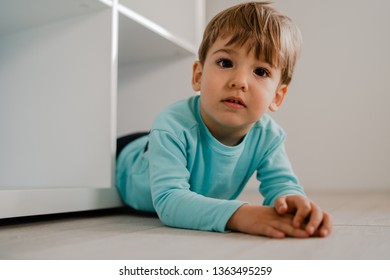 Portrait Of A Little Boy In Blue At Home Lying In The Book Shelf On The Floor Hiding Games