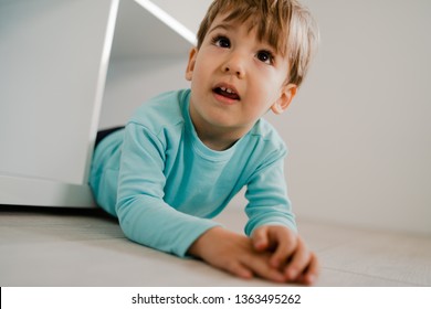 Portrait Of A Little Boy In Blue At Home Lying In The Book Shelf On The Floor Hiding Games