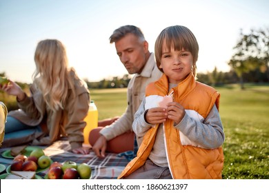 Portrait Of Little Boy Eating Sandwich And Looking At Camera With A Smile. Young Parents Having Picnic With Their Children On Blanket In Park. Family And Kids, Nature Concept. Horizontal Shot