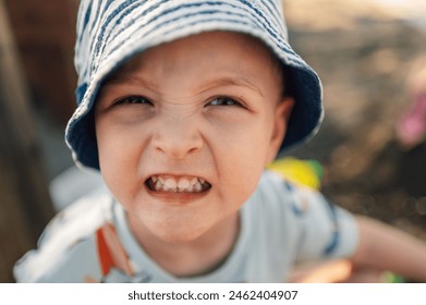 Portrait of a little boy frowning and being angry. Portrait of a child with cap frowning and looking at camera while spending day outdoors. An angry cute little boy is sitting outdoors and frowning. - Powered by Shutterstock