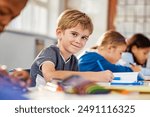 Portrait of little boy studying in classroom at school. Young happy caucasian boy doing his elementary school homework while sitting at desk with his classmates. Elementary student looking at camera.