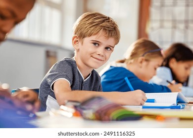 Portrait of little boy studying in classroom at school. Young happy caucasian boy doing his elementary school homework while sitting at desk with his classmates. Elementary student looking at camera.