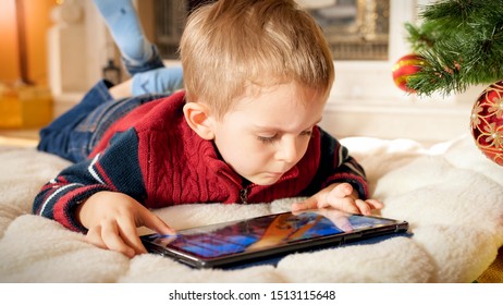 Portrait Of Little Boy With Tablet Lying On Floor Under Christmas Tree On NEw Years Morning