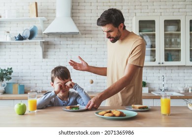 Portrait of a little boy who does not want to eat and his angry dad makes him eat sitting at the table during breakfast at home in the kitchen.