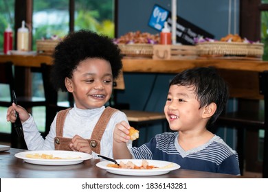 Portrait Of Little Cute Black Boy And Asian Boy Eating French Fries Potato With Ketchup In The Cafe. African American Kid And White Kid Enjoy Eating French Fries. Happy Child Eating Unhealthy Food.