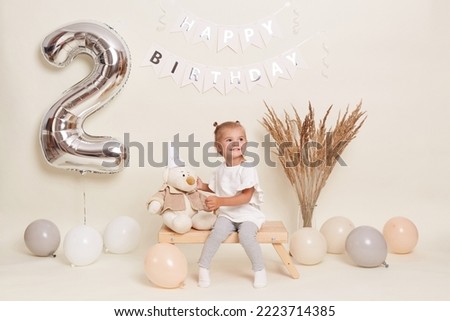 Portrait of little cute blond girl sitting on wooden bench among balloons, celebrating two years, posing in festive room with decoration and big number two made of inflatable foil balloon.