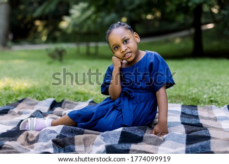 Portrait of little cute happy African girl in blue dress, sitting on checkered blanket in summer park, having a picnic time. Happu childhood, summer portrait, copy space