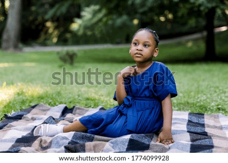 Portrait of little cute happy African girl in blue dress, sitting on checkered blanket in summer park, having a picnic time. Happu childhood, summer portrait, copy space