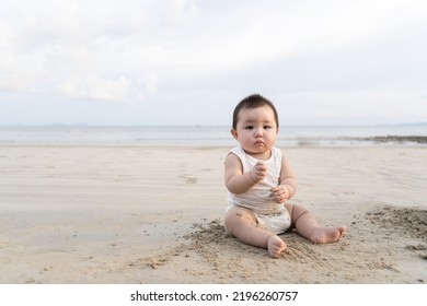 Portrait Of A Little Cute Toddler Asian Girl Wearing White Bodysuit Sitting During Summer Vacations. Baby Girl Playing In The Sandy Beach.