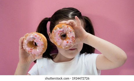 Portrait of little dark hair girl peeping through donut with icing in her hands, look at the camera. Pink background.