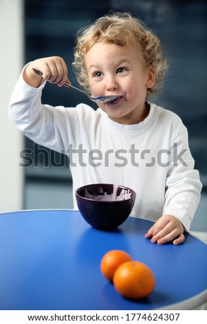 Portrait of the little funny, cheerful boy having a meal. Cute child in a white shirt with a big spoon having breakfast, lunch or dinner. 