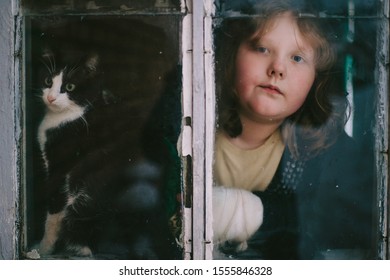 Portrait of little funny girl with her lovely kitten looking out of old window in village house.