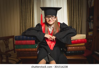 Portrait Of Little Genius Girl In Graduation Clothes Leaning On Books At Library