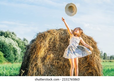 Portrait of little girl in blue dress catching hat, jumping near haystack in field. Light sunny cloudy day. Joy and glad concept. Blue clear sky on background. Wind taking away straw hat. Low angle - Shutterstock ID 2151765863