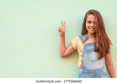 Portrait Of A Little Girl Child In A Denim Jumpsuit 9-13 Years Old On An Empty Wall, The Girl Shows Two Fingers Symbolizing That Everything Is Good