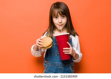 Portrait Of A Little Girl Dressed Casually And Taking A Sip Of A Large Soda While Holding A Burger In The Other Hand