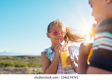 Portrait of little girl drinking orange juice in a glass with straw. Rear view of brother drinking with sister at outdoor park. Boy and cute girl with sunglasses suck from the straw a fresh juice.  - Powered by Shutterstock