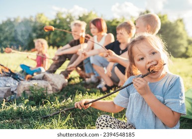 Portrait of little girl eating grilled sausage. Group of Kids - Boys and girls roasting sausages on long sticks over a campfire flame. Outdoor active time spending or camping in Nature concept.  - Powered by Shutterstock