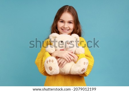 Portrait of little girl hugging cute white toy bear, looking at camera, dreaming, enjoying present, wearing yellow casual style sweater. Indoor studio shot isolated on blue background.