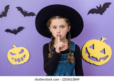 Portrait Of Little Girl Kid In Big Hat Say Hush Be Quiet With Finger On Lips Shhh Gesture, Posing Isolated On Purple Background Studio Decorated Yellow Funny Pumpkins And Black Bats. Halloween Concept