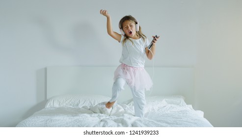 Portrait Of A Little Girl Listening To The Music With The Headphones And Dancing On Her Parents Bed. Concept: Music, Freedom, Happiness