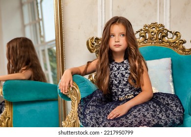 Portrait Little Girl Princess 5-6 Years Old, Fashion Model In Stylish Elegant Blue Dress Sitting On Armchair In Studio With Mirror, Thinking, Looking Away. Fashionable Lady Actress At Home. Copy Space