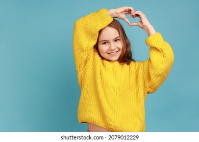 Portrait of little girl shows heart shape with fingers above head, expresses innocent childhood love, wearing yellow casual style sweater. Indoor studio shot isolated on blue background.