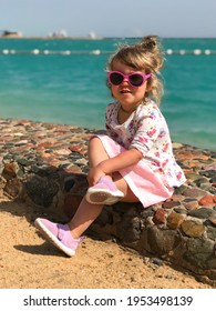 Portrait Of A Little Girl Sitting On A Stone Embankment In Sunglasses Against The Backdrop Of A Seascape. Concept:a Little Fashionista Is Relaxing On The Beach, A Photo Shoot For Tourists.