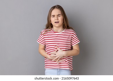 Portrait Of Little Girl Wearing Striped T-shirt Clutching Belly, Feeling Discomfort Or Pain In Stomach, Sick Child Suffering Constipation Cramps. Indoor Studio Shot Isolated On Gray Background.